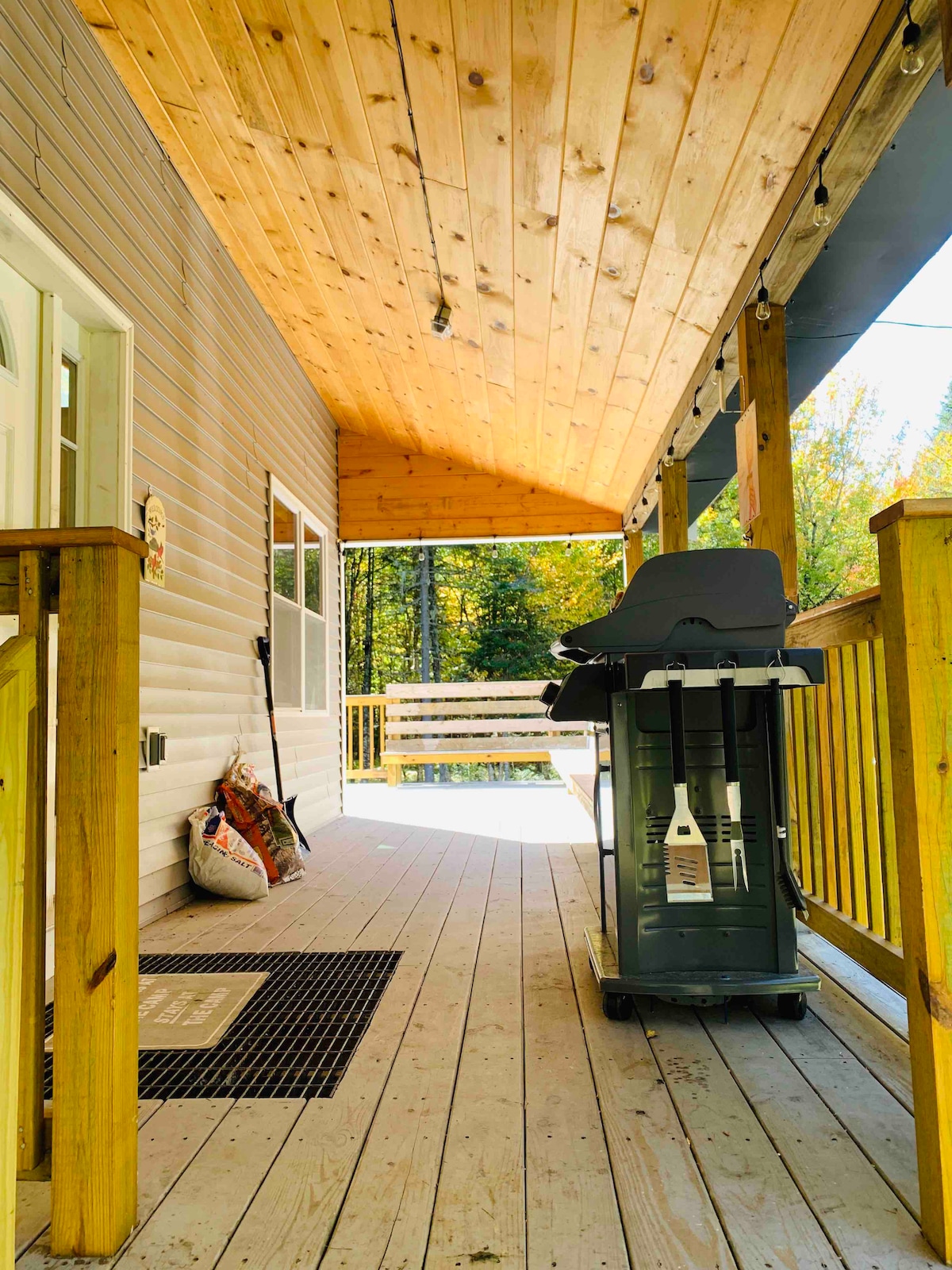 A covered outdoor deck is visible, featuring wooden flooring and warm wooden beams overhead. A grill is positioned beside a welcome mat, and nearby, a seating area can be seen surrounded by trees. Soft lighting strings hang along the ceiling, adding a warm touch.