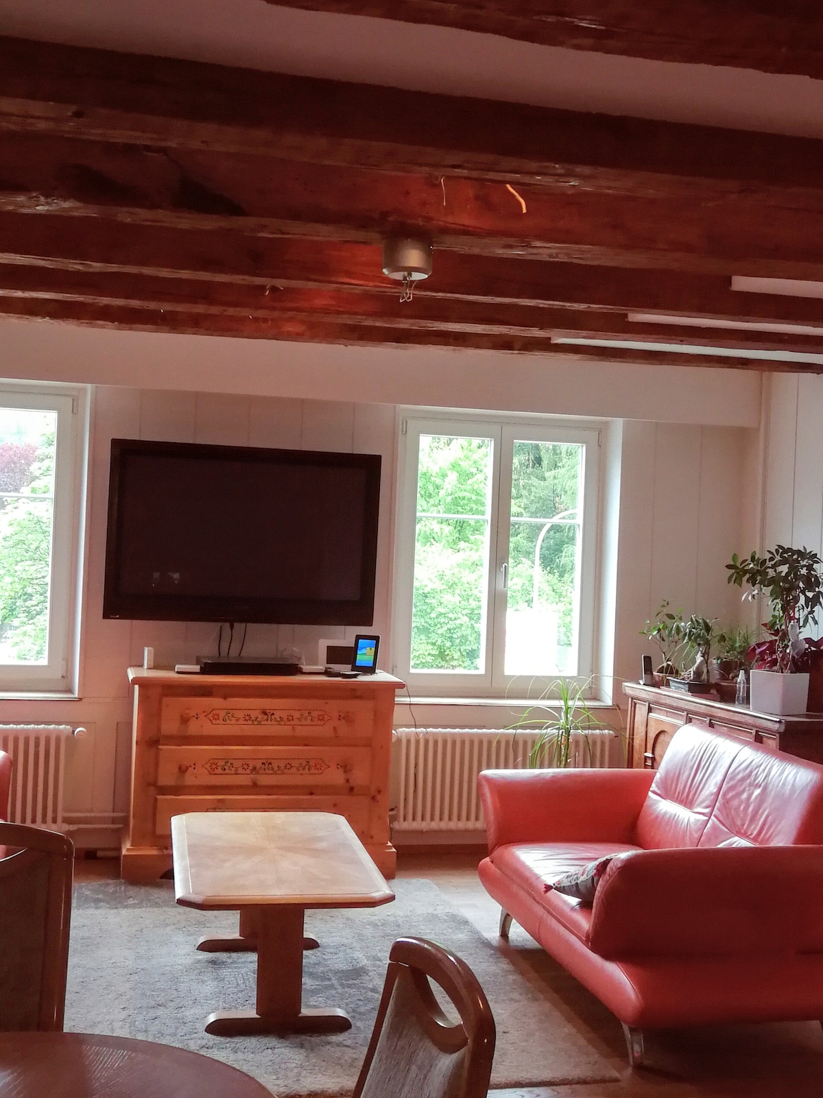 A living area features wooden beams overhead, with a television mounted on a wall. Two large windows allow natural light to illuminate the space, framed by greenery outside. A wooden table is situated in front of a pair of red sofas, enhancing the cozy atmosphere.