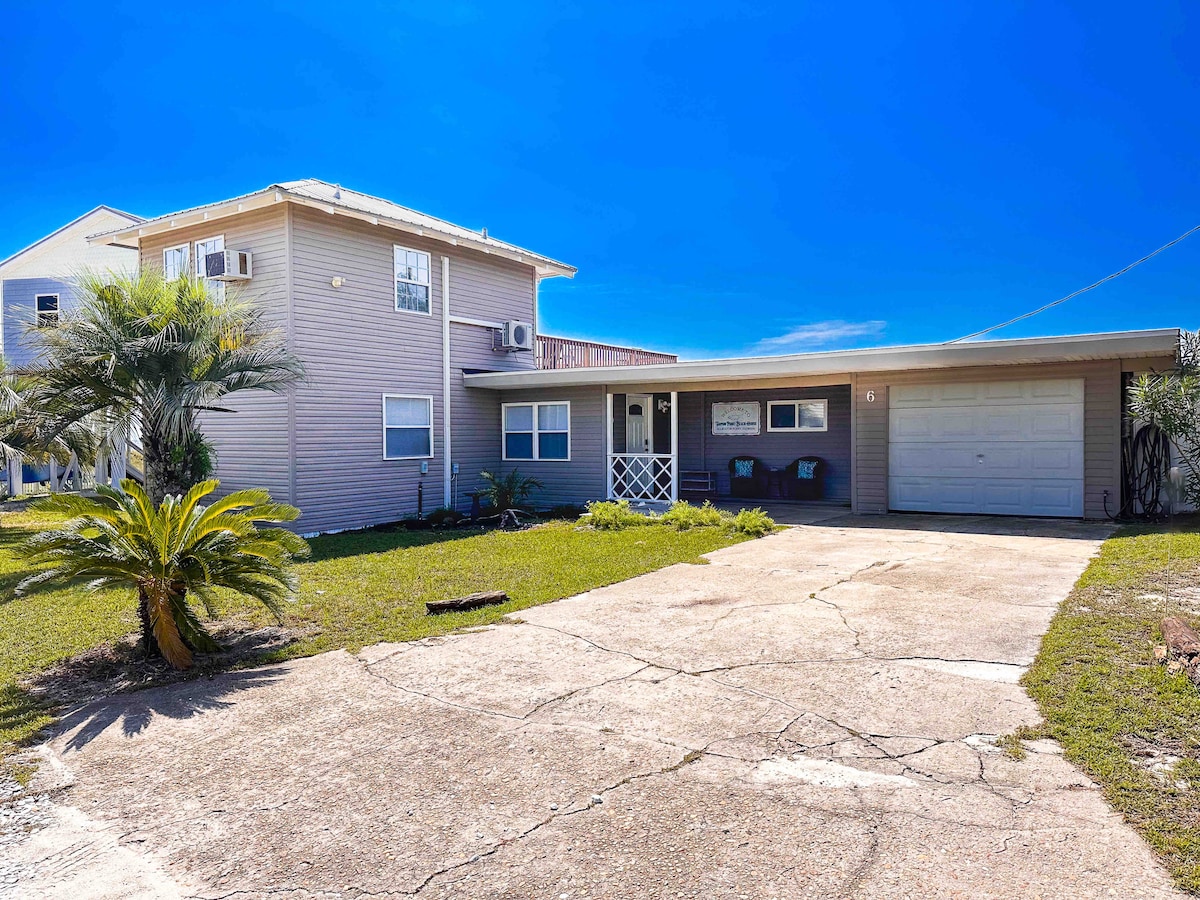 A two-story beach bungalow is depicted with pale siding, featuring a wide driveway leading up to the front entrance. The surrounding greenery includes palm trees, enhancing the coastal vibe. A garage is visible on the right side, with a clear blue sky providing a bright backdrop.