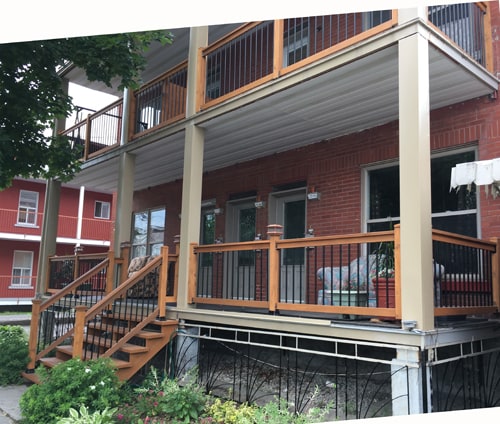 The entrance features a set of wooden stairs leading up to a covered porch with multiple seating areas and a railing. The brick facade of the building is complemented by greenery in the foreground, providing a welcoming approach to the property.