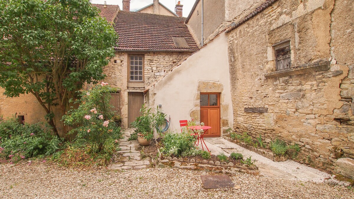 A charming outdoor area is showcased, featuring a small patio with a round table and two red chairs. Surrounding greenery and a stone wall exhibit natural textures. The entrance to the house is visible, along with a staircase leading up to the upper level.