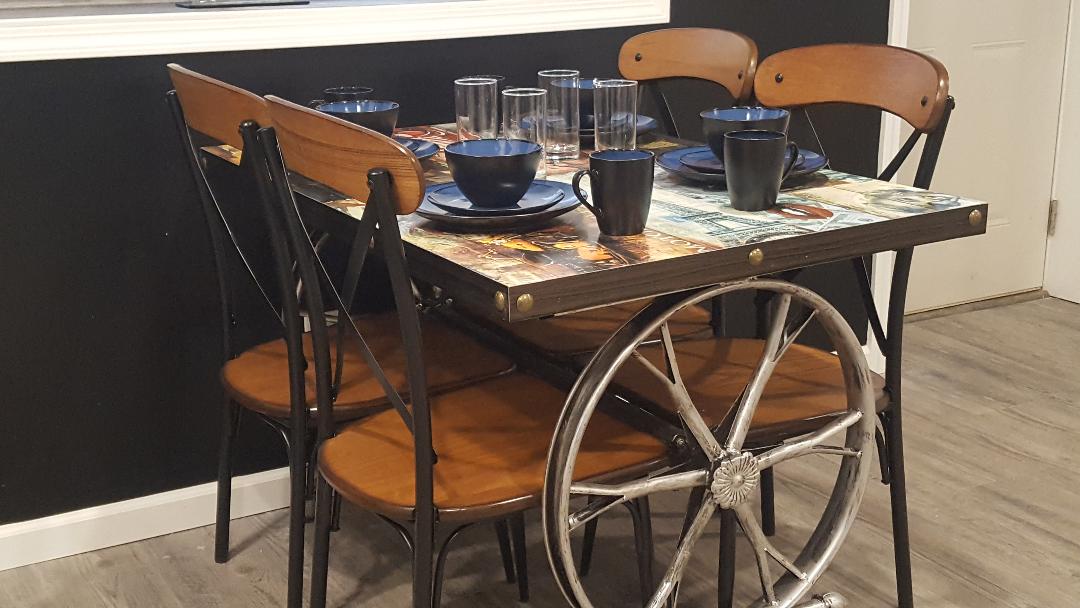 A dining table with a unique cart wheel base features four wooden chairs. The tabletop displays an artistic design, and is set with four bowls and cups. Glasses are neatly arranged nearby, offering a functional and inviting dining space.