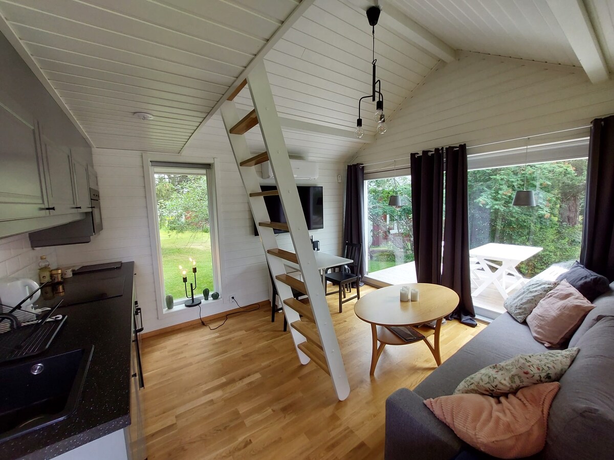 An inviting interior features a cozy seating area with a grey sofa. A circular coffee table is positioned in front, while a multi-step staircase leads to the sleeping loft. Large windows allow natural light to fill the space, offering views of the surrounding greenery.