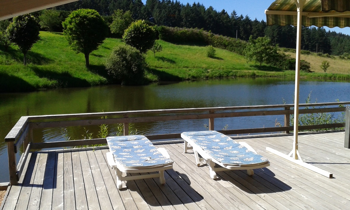 A wooden deck features two sun loungers positioned toward a tranquil pond surrounded by lush greenery. A parasol provides shade, and the serene landscape includes rolling hills and trees in the background.