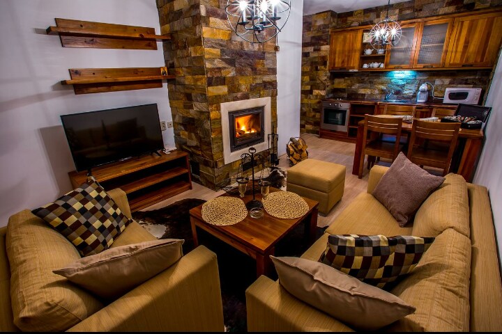 A cozy living space features a stone fireplace surrounded by wooden shelves and a flat-screen TV. Comfortable seating is provided on beige couches adorned with patterned cushions. A wooden dining area can be seen in the background, with natural light illuminating the warm tones of the space.