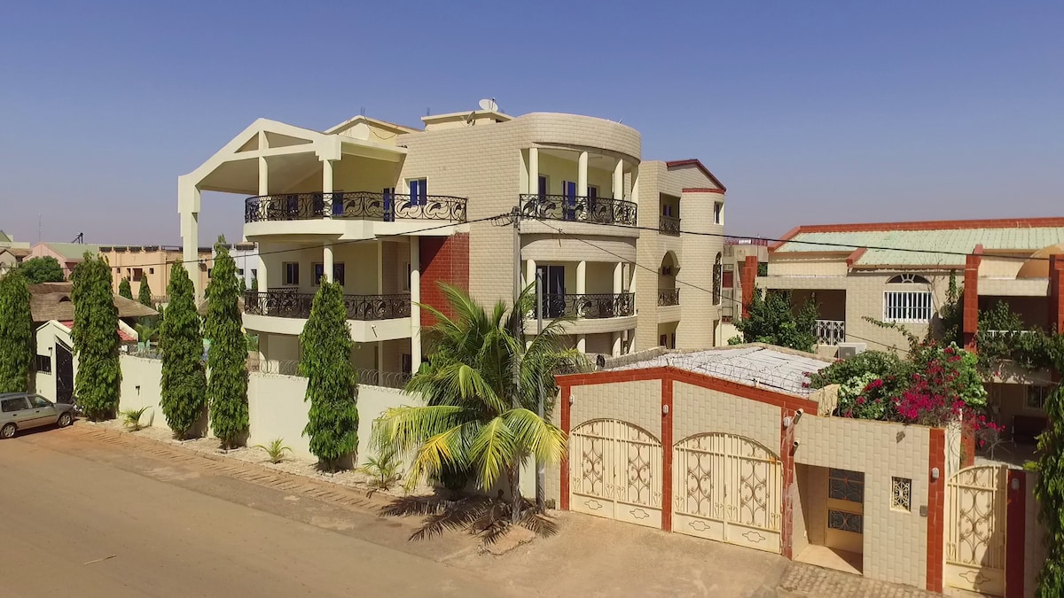 The exterior of the residence is showcased, featuring a modern three-story building with multiple balconies and a variety of greenery. A wrought iron gate provides entrance, while the surrounding area includes paved pathways and nearby vehicles, emphasizing a welcoming atmosphere.