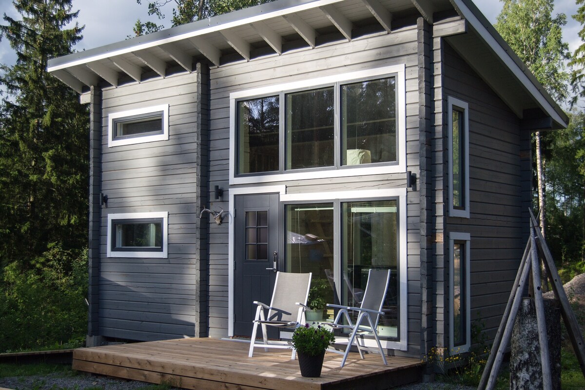 A modern cabin exterior is presented with large windows that allow natural light to flood the interior. A wooden deck features two lounge chairs, inviting relaxation. Surrounding greenery enhances the serene outdoor environment.