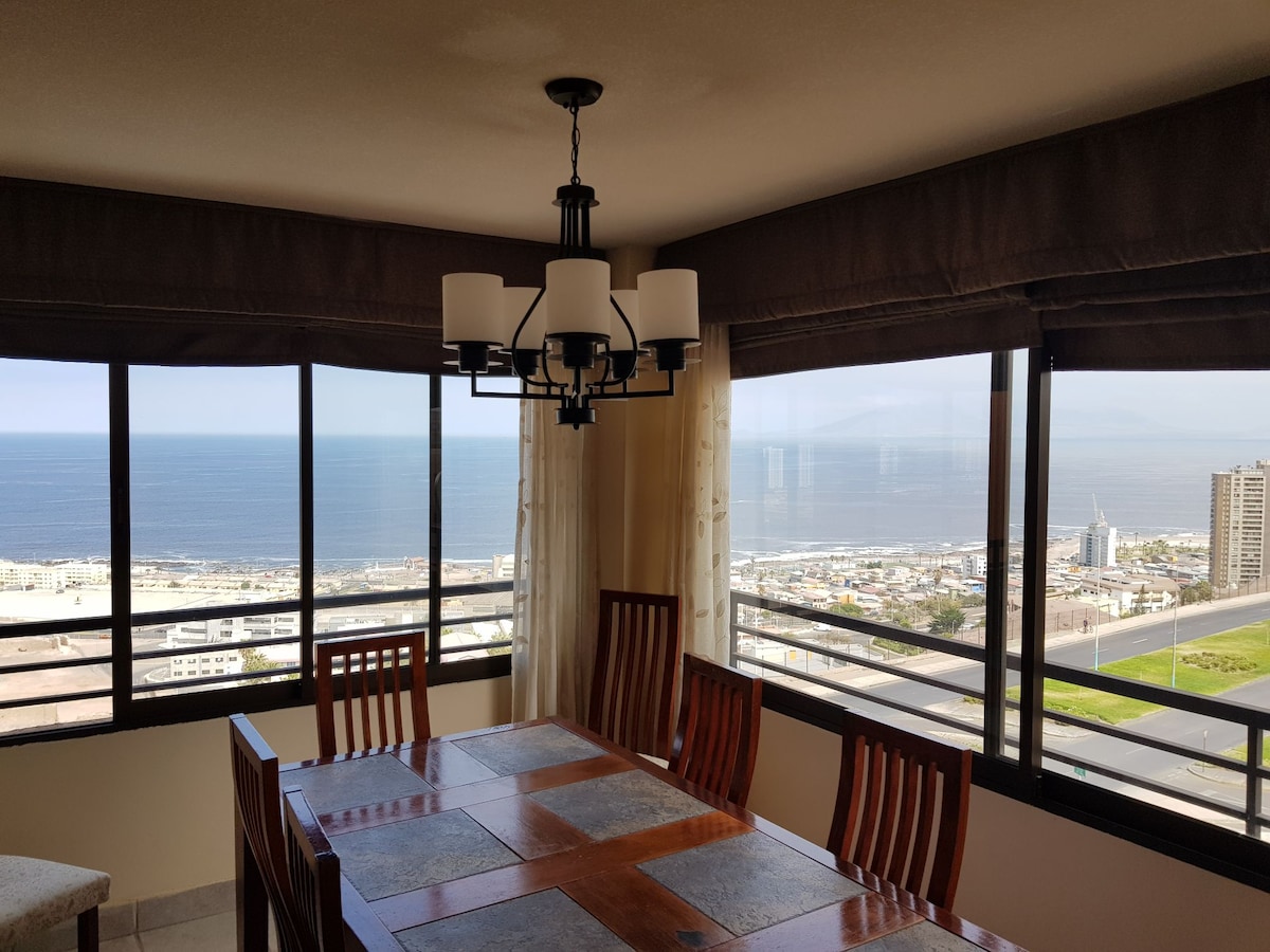 A dining area features a wooden table surrounded by several chairs, positioned near large windows offering expansive ocean views. Natural light filters through sheer curtains, accentuating the spaciousness of the room while highlighting the coastal scenery beyond.