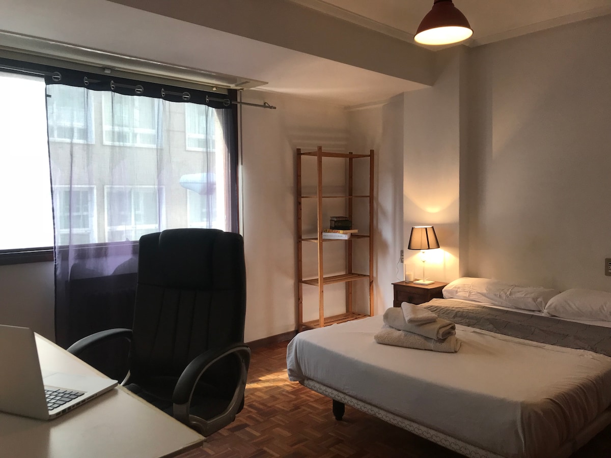 A well-lit bedroom features a spacious bed with neatly arranged linens. A comfortable desk chair is positioned beside a desk equipped with a laptop. A wooden shelving unit stands against the wall, complemented by a small lamp that emits soft lighting near the bed.