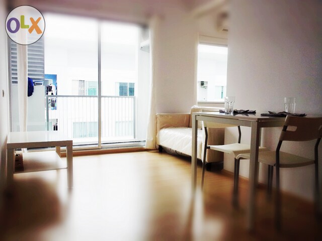 A bright and airy living space features large sliding glass doors leading to a balcony. A small dining table with two chairs sits adjacent to a sofa bed, with a coffee table positioned nearby. Natural light fills the room, creating a welcoming environment.