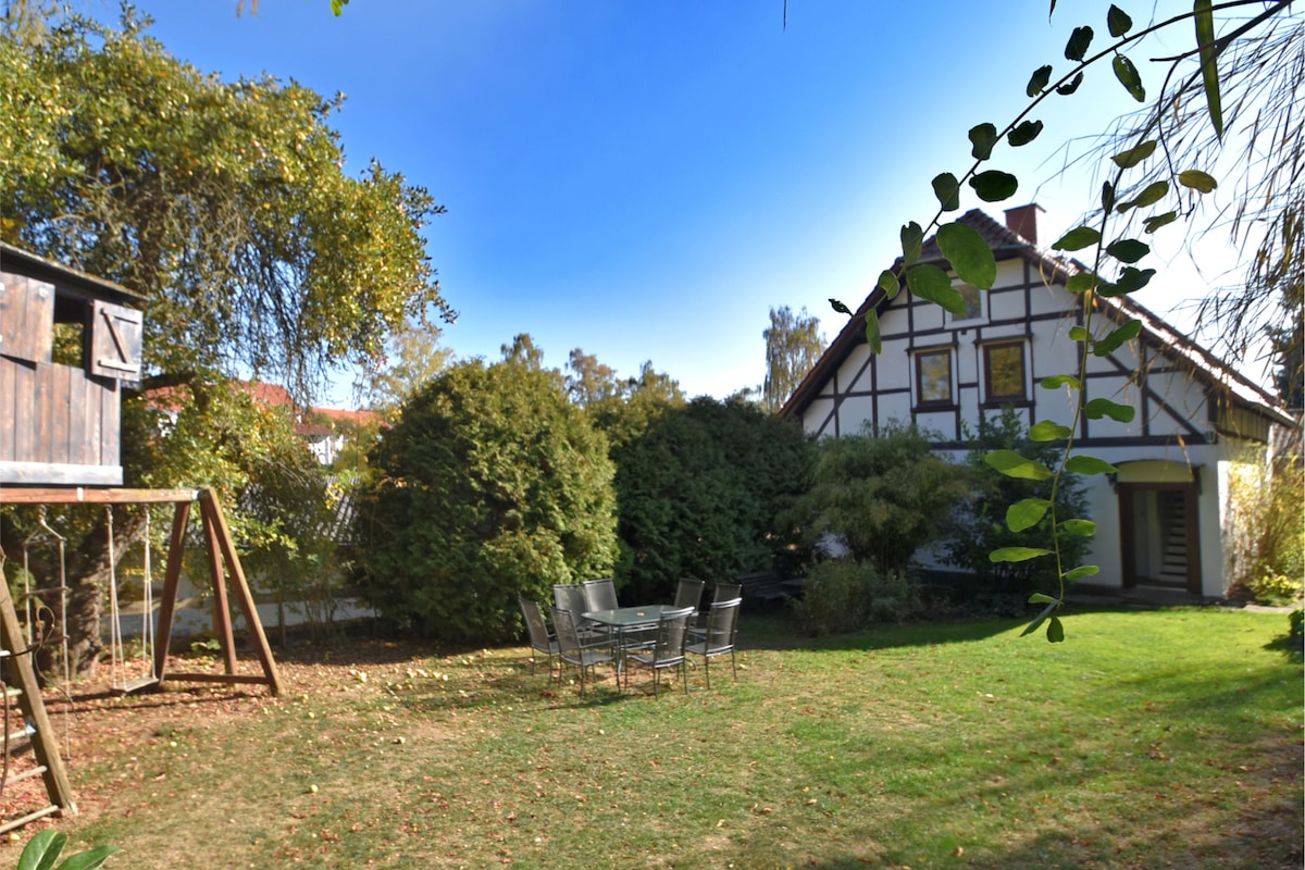 A spacious garden is depicted, featuring well-maintained grass and a mix of trees providing privacy. A wooden tree house is seen alongside a swing set. A large outdoor dining table surrounded by metal chairs is positioned on the grass, ready for gatherings.