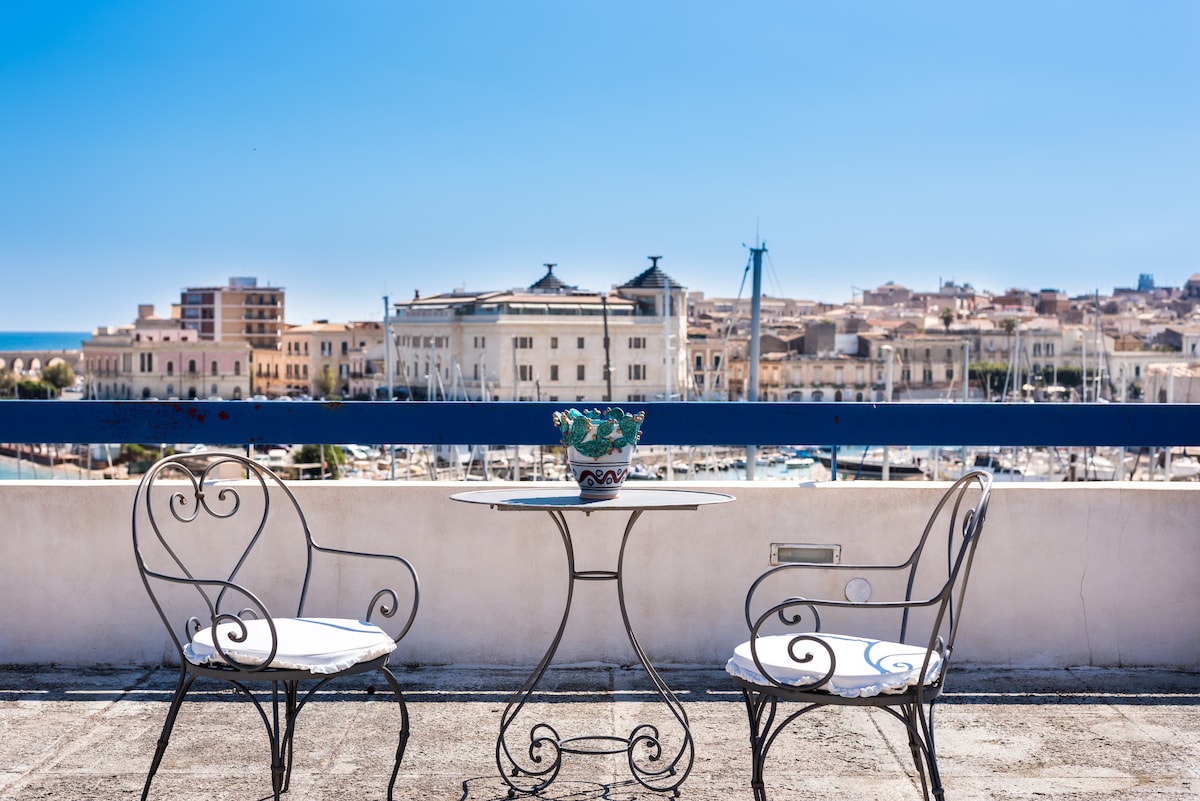 A small patio area is visible, featuring a round table accompanied by two wrought-iron chairs. A colorful potted plant rests at the center of the table. The expansive view of the city and marina can be seen in the background under clear blue skies.