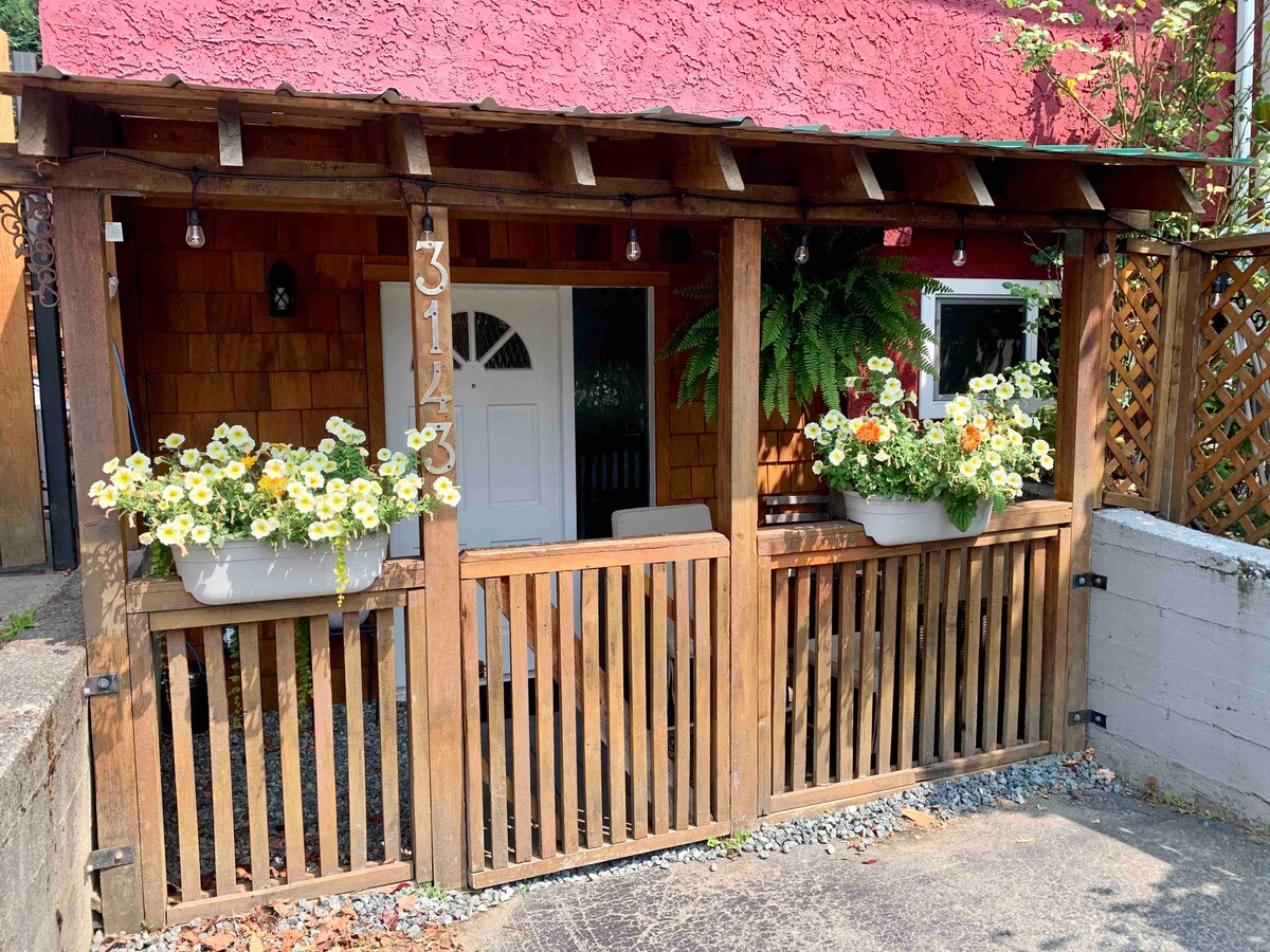 The exterior entrance features a welcoming porch with wooden railings and lush planters filled with bright flowers. A cozy seating area is visible behind the door, which is set in a charming, rustic wooden structure adorned with natural elements.