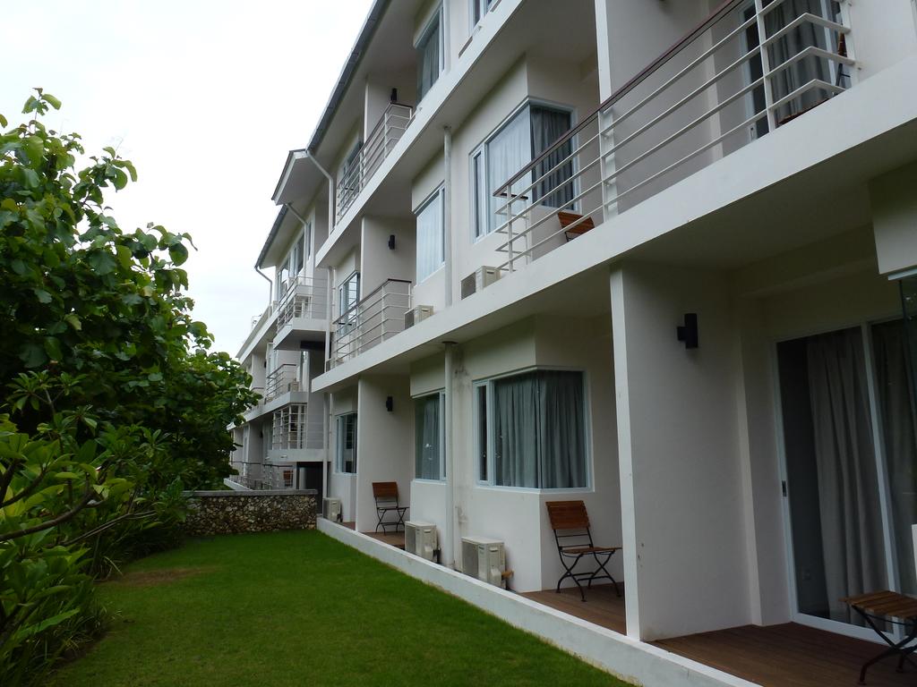 A modern building features multiple balconies equipped with wooden chairs, surrounded by manicured greenery. Large windows allow natural light into the interior, while air conditioning units are discreetly placed below each window. The peaceful environment offers a sense of tranquility.