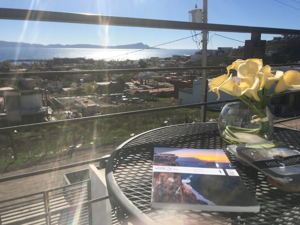 A balcony table is set with a magazine and a small vase of yellow flowers. The view captures the ocean and distant hills under clear blue skies, with urban and natural landscapes visible in the background.