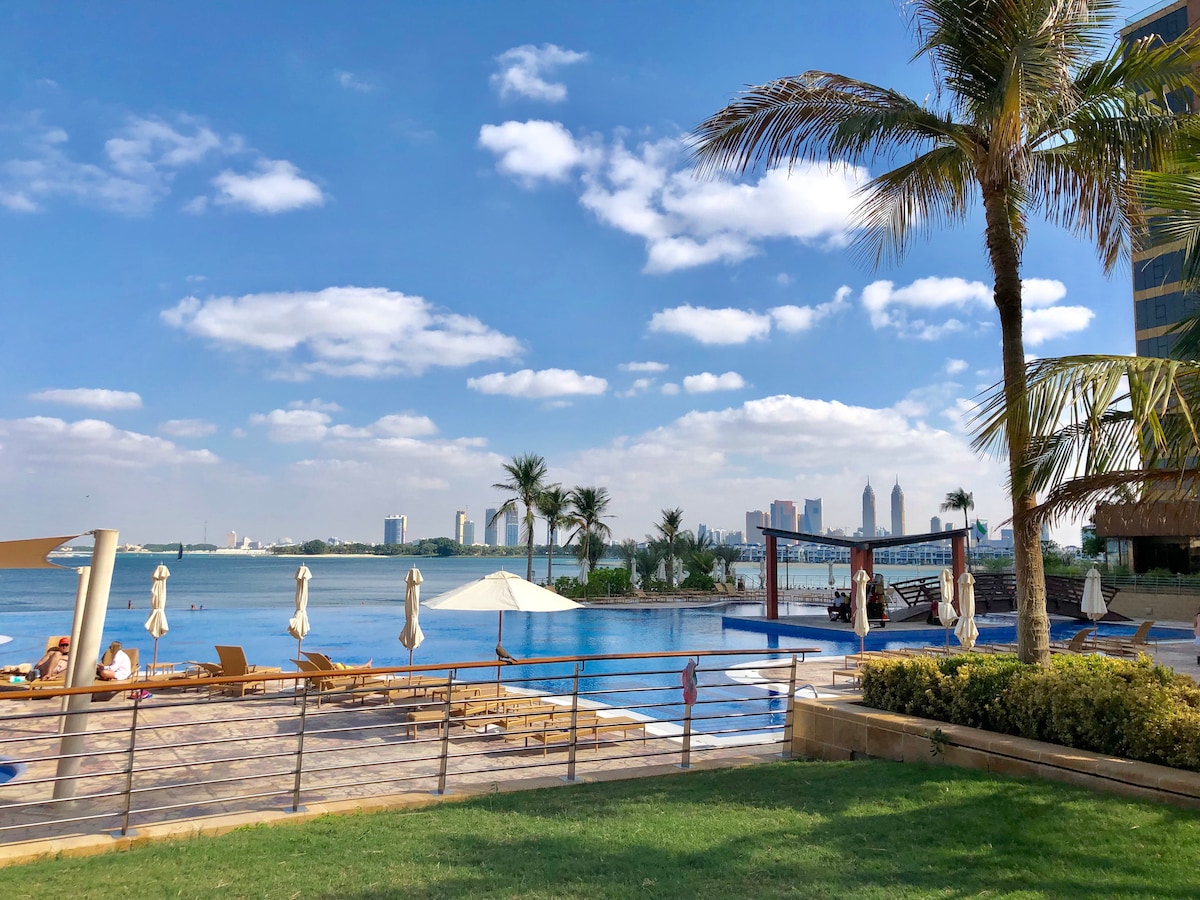 An expansive infinity pool is visible, surrounded by sun loungers and umbrellas. Lush greenery lines the area, with palm trees adding a tropical feel. The backdrop features the shimmering Arabian Sea and city skyline under a bright blue sky adorned with fluffy clouds.