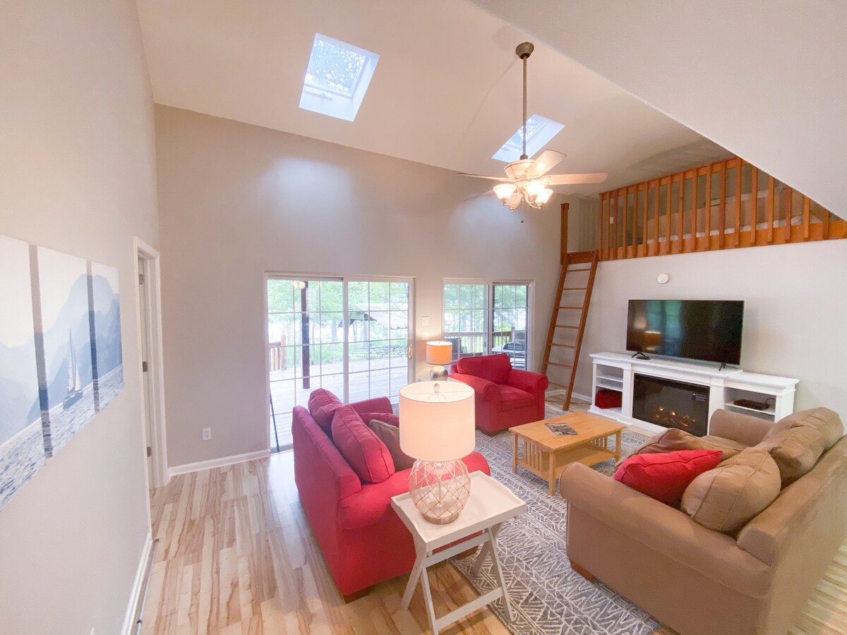 A bright living area is captured from above, featuring comfortable seating with two sofas and a coffee table. Large glass doors provide access to a deck, while a flat-screen TV rests on an entertainment unit. Natural light streams in through skylights.