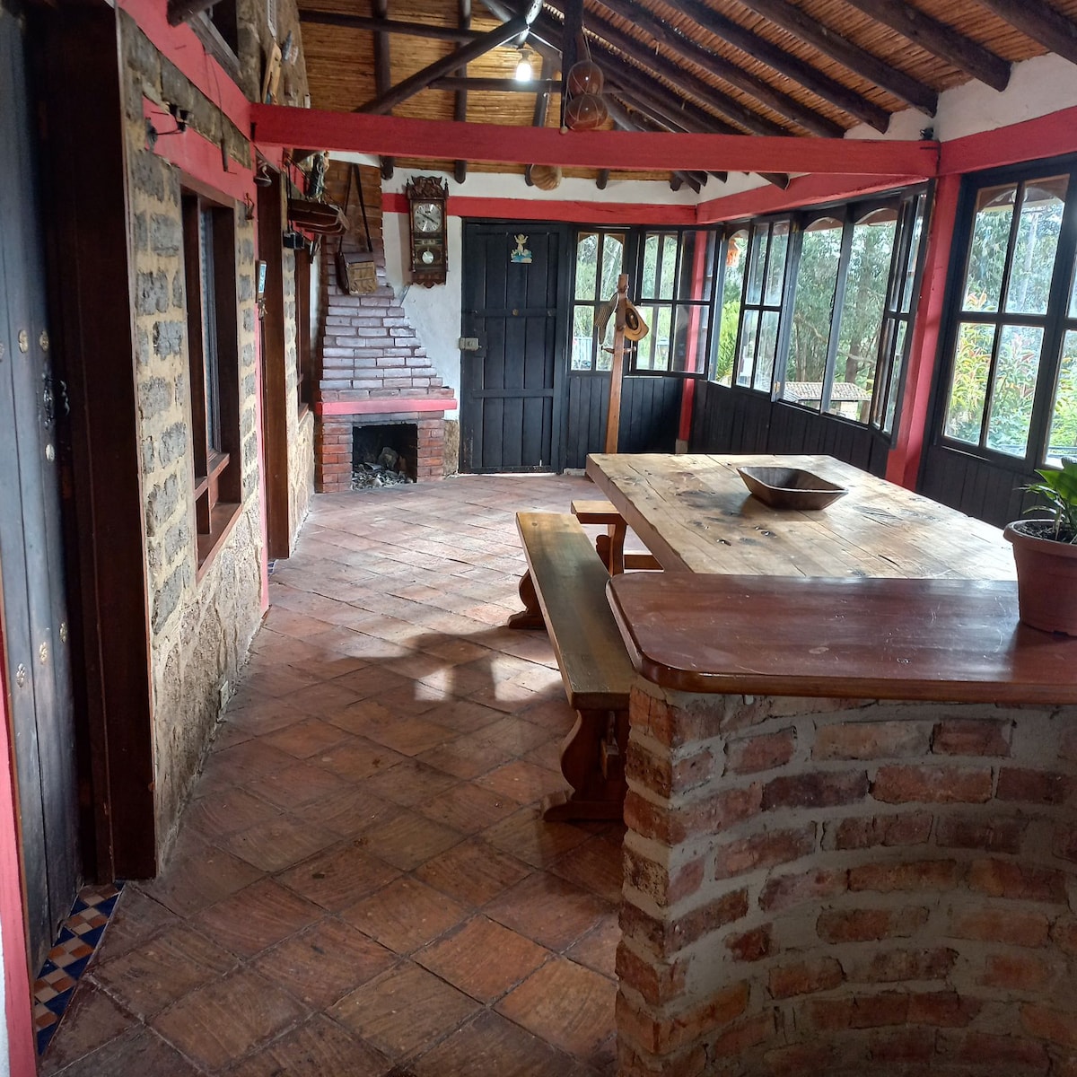 A warm and inviting common area features a large wooden table surrounded by benches, set against rustic stone walls. Large windows allow natural light to fill the space, while a cozy fireplace adds to the atmosphere. Exposed wooden beams and red accents enhance the charm.