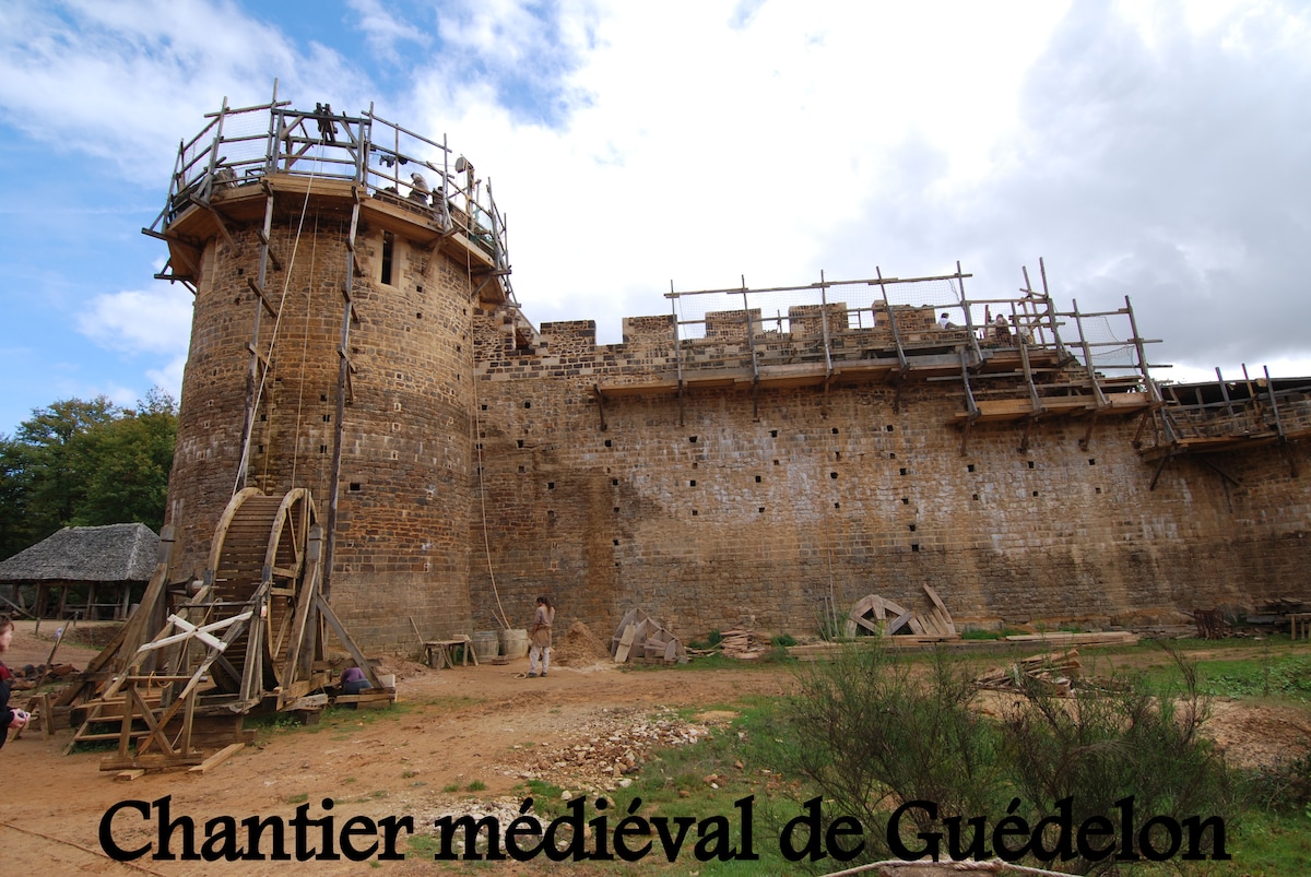 A medieval construction site is displayed, featuring a stone wall structure under construction with scaffolding. Nearby, a large wooden wheel is visible, alongside various tools and materials used in the building process. Surrounding greenery adds a natural backdrop.