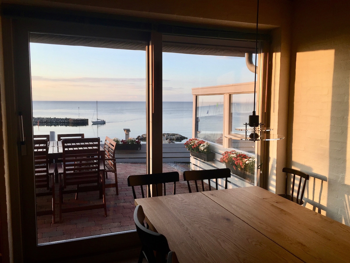 A dining space is highlighted by a large wooden table, surrounded by black chairs. Expansive sliding glass doors provide a view of the sea and outdoor seating area, with colorful flowers visible on the terrace. Natural light filters into the room, creating a serene atmosphere.