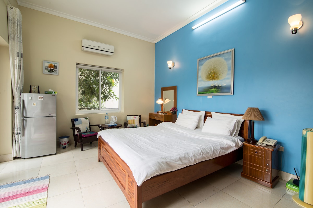 A spacious bedroom is highlighted by a large wooden bed with white bedding. A blue accent wall provides a pop of color. Natural light filters through the window, and a small seating area with dark chairs and a lamp is present. A mini fridge is visible in the corner.