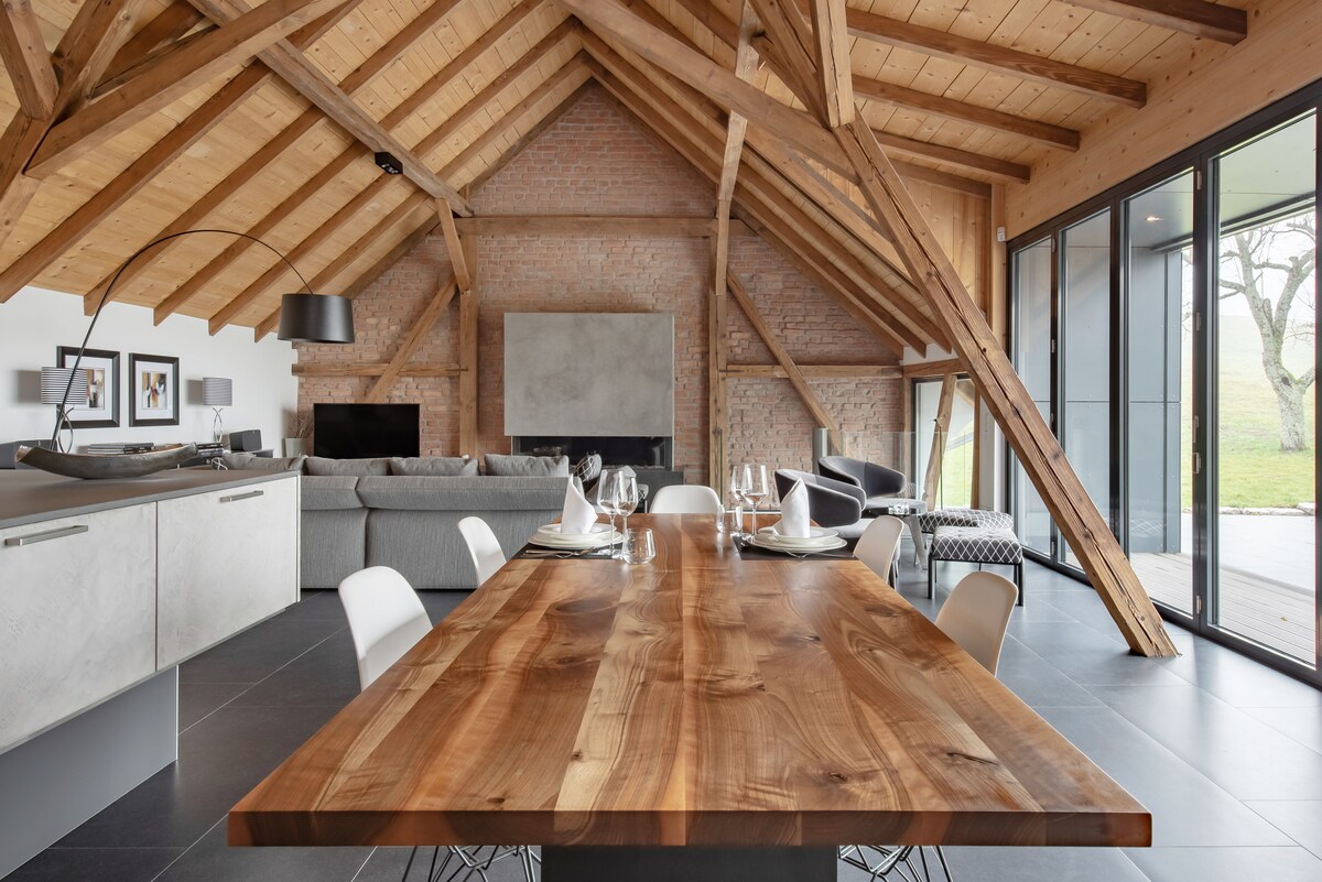 A spacious living and dining area is highlighted by a large wooden table set for dining. High ceilings with exposed beams create an airy feel, complemented by a cozy seating area and large glass doors leading to the terrace, offering picturesque views of the surrounding nature.