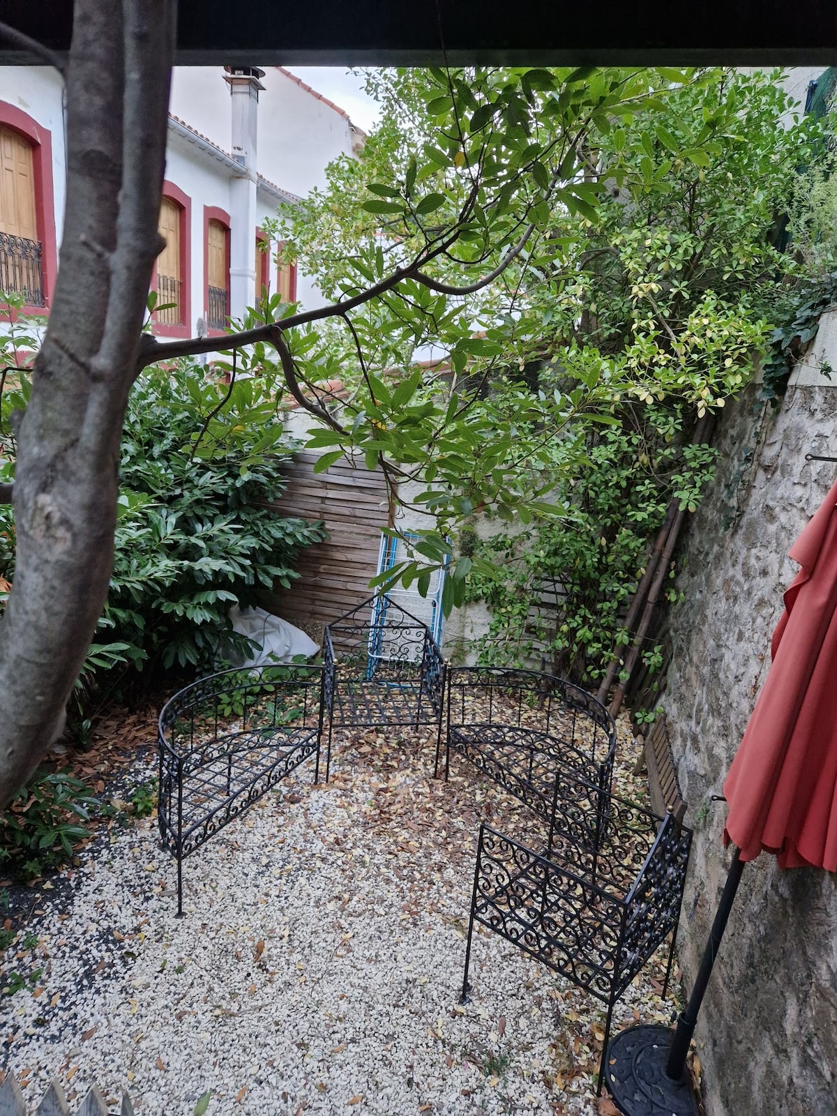 A tranquil garden area is enclosed by a decorative metal fence, featuring a gravel surface scattered with leaves. Lush greenery and a tree provide natural shade, while a red patio umbrella stands nearby, enhancing the calm atmosphere of the space.