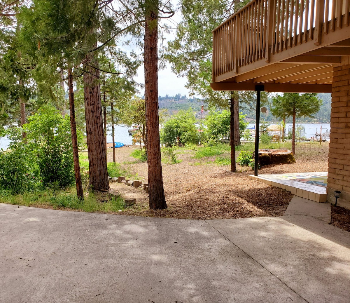 A serene outdoor space is depicted, featuring tall pine trees and a gravel pathway leading towards the lake. A patio area is seen in the foreground, with glimpses of lush greenery and the water beyond, creating a peaceful atmosphere ideal for relaxation.