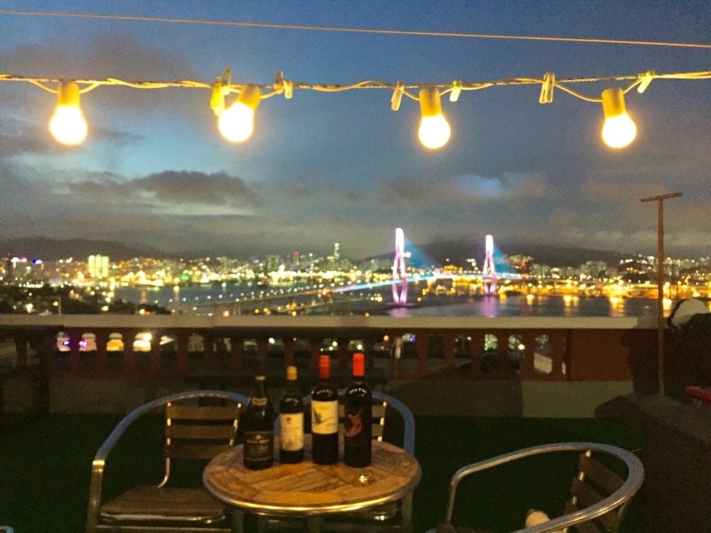 The rooftop area is illuminated by string lights, creating a warm ambiance. A wooden table is surrounded by metal chairs and a selection of wine bottles lines the table. The skyline of Busan is in the background, showcasing the illuminated city and the harbor bridge.