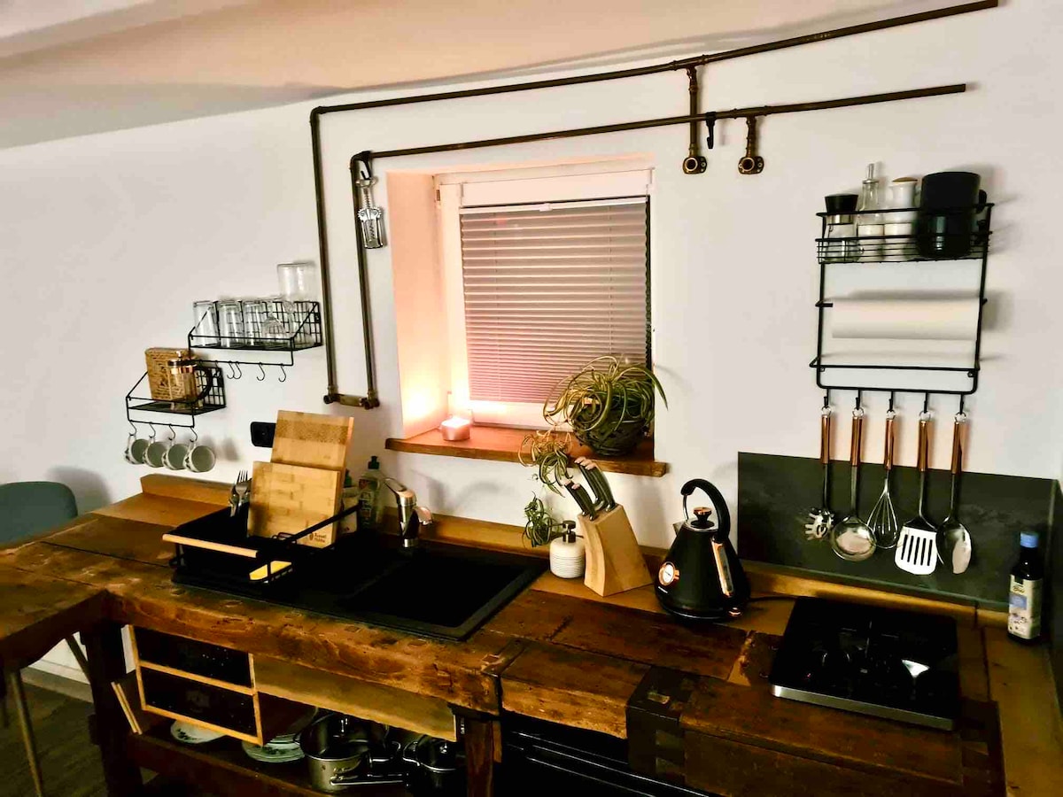 A rustic kitchen area is highlighted by a wooden workbench converted into a functional countertop. Essential kitchen tools and utensils are neatly organized on wall-mounted racks. A window allows natural light to enter, while a small candle adds a touch of warmth to the space.