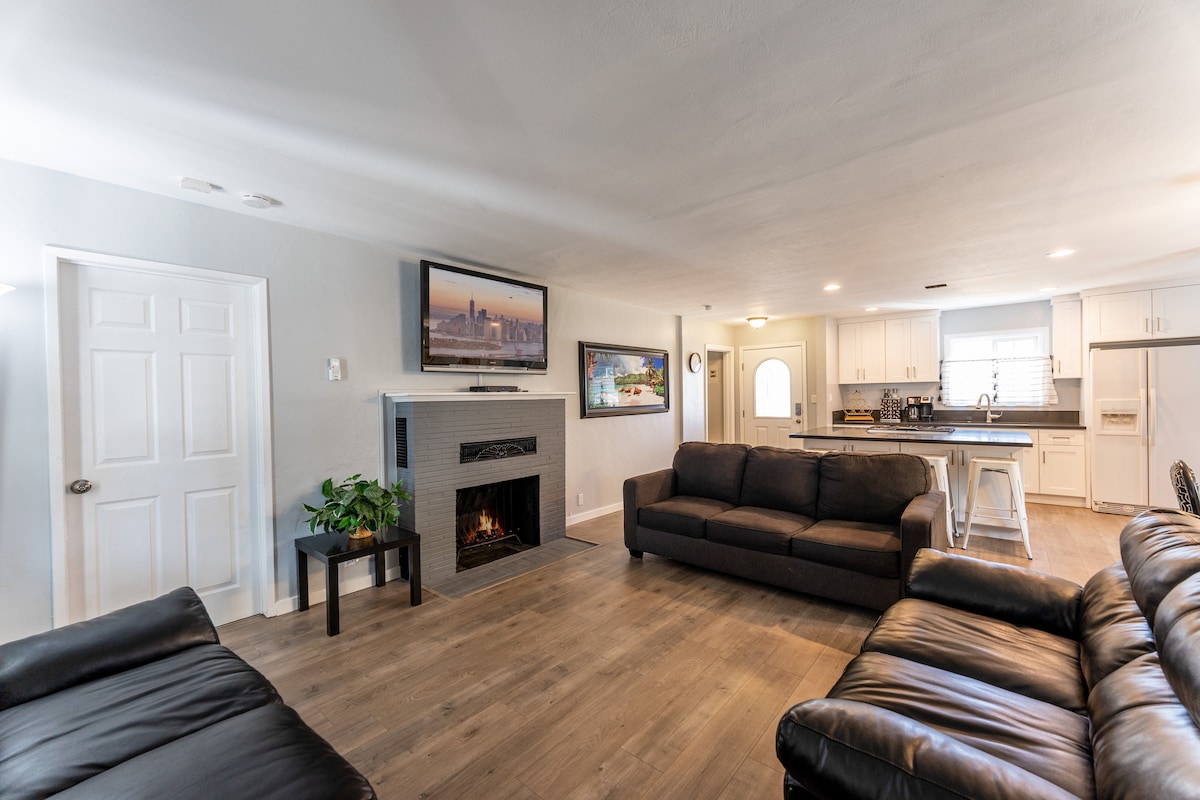 A spacious living area features two sets of comfortable couches arranged around a modern fireplace. A flat-screen TV is mounted above the fireplace, and a doorway leads to a bright kitchen that is partially visible. Light-colored walls give the room a welcoming feel.