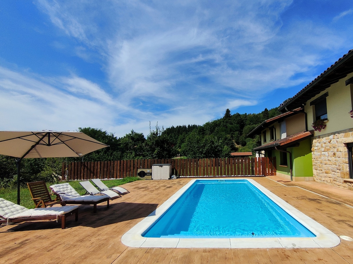A spacious outdoor area features a rectangular swimming pool surrounded by a wooden deck. Several lounge chairs are positioned alongside the pool, with an umbrella providing shade. Lush greenery and a wooden fence create a natural backdrop, enhancing the inviting space.
