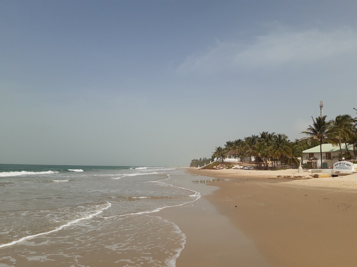A sandy beach is shown with gentle waves lapping at the shore. Palm trees are visible along the coast, providing shade and greenery. A few buildings can be seen in the distance, set against a clear blue sky.