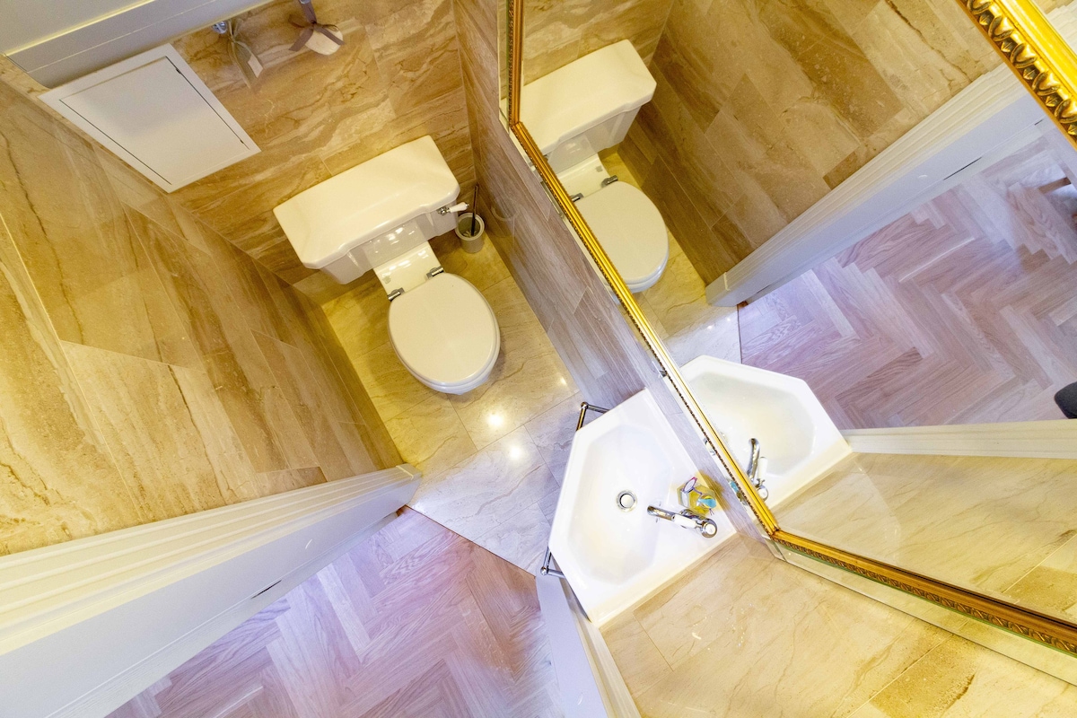 A spacious bathroom features modern fixtures, including a toilet and sink. Elegant marble flooring reflects light, enhancing the room's brightness. Mirrors extend across the walls, creating a sense of depth. The overall design promotes a clean and fresh environment.