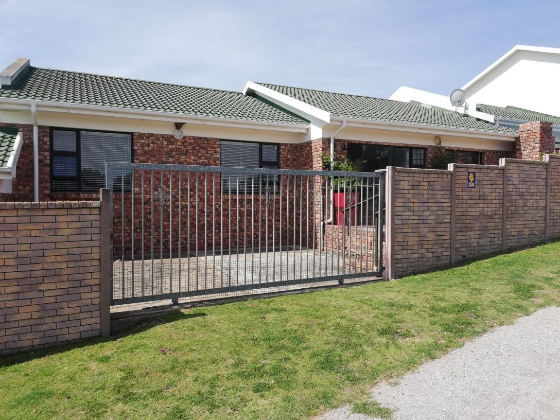A single-story holiday house is displayed behind a low brick wall and a gated entrance. The structure features a combination of brick and white siding, with a green tiled roof. Sunlight highlights the landscaped front yard with well-maintained grass.