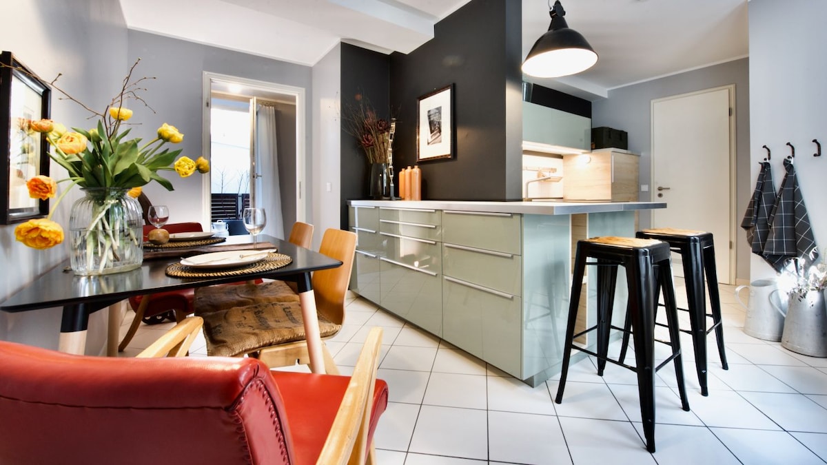 A modern kitchen and dining area is showcased, featuring a large central island with storage drawers. Wooden dining chairs surround a black table set for meals. Colorful flowers in a vase add a touch of vibrance, while sleek bar stools enhance the contemporary atmosphere.