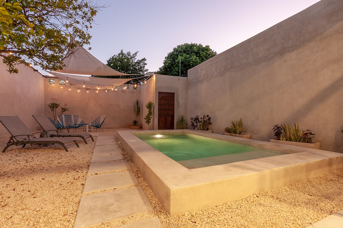 An outdoor area features a private swimming pool surrounded by a smooth stone deck. Lounge chairs are positioned nearby, and string lights hang above, providing a soft glow. Lush plants line the walls, creating a serene ambiance during twilight.