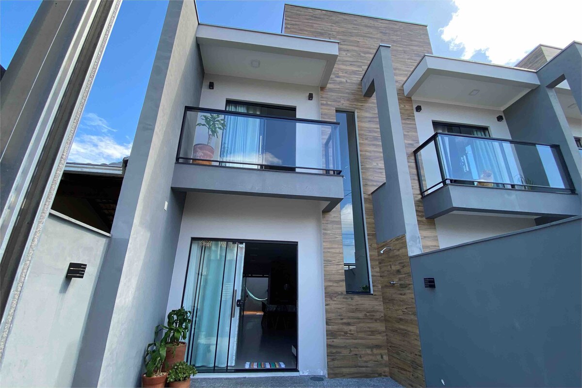 The exterior of a modern two-story house is presented, featuring a combination of light-colored walls and wooden accents. Glass balconies on the upper level enhance the design. Potted plants are seen near the entrance, with a large glass door leading into the interior.