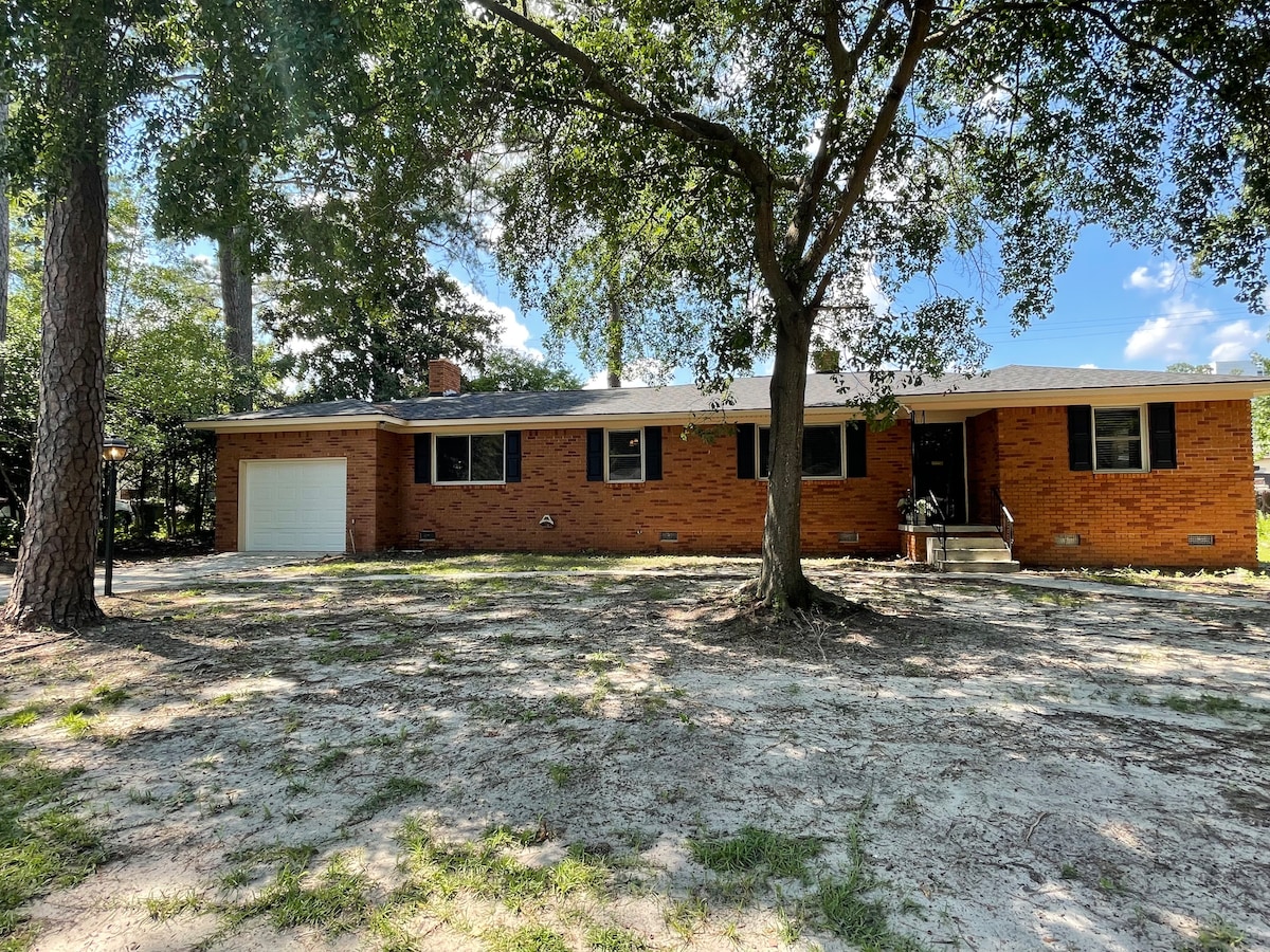 The exterior of the home showcases a brick façade complemented by a manicured yard. A single garage and a pathway lead to the entrance, surrounded by mature trees, with dappled sunlight filtering through the leaves and a clear blue sky overhead.
