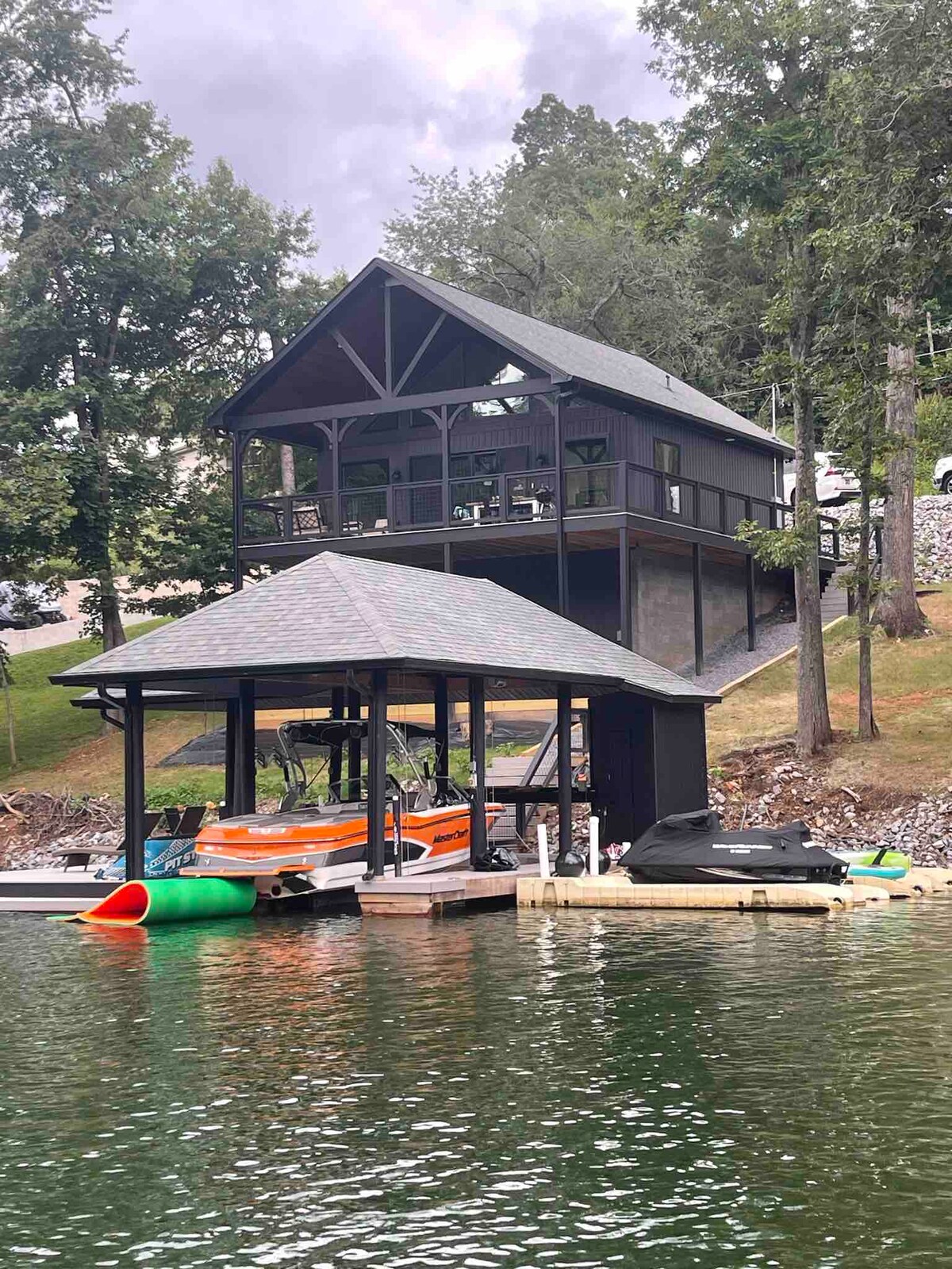 A two-story lakefront property is prominently displayed, featuring a covered dock below. The structure is dark in color, maintaining a rustic appearance amidst surrounding greenery. Boats can be seen moored at the dock, reflecting on the water's surface.