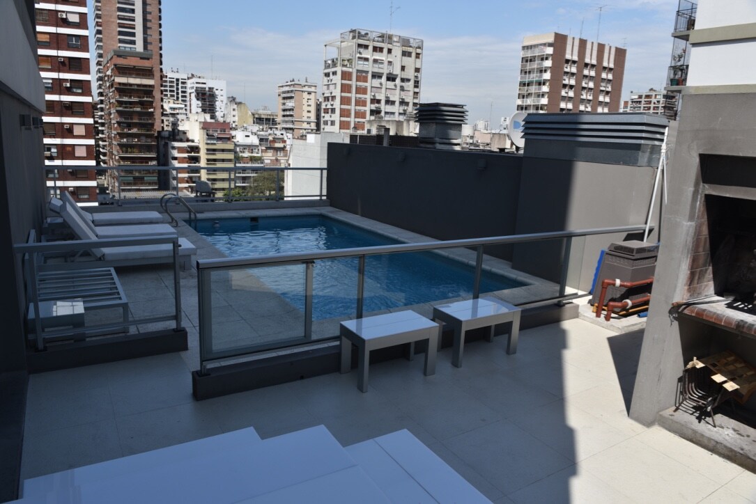 A rooftop area features a swimming pool surrounded by a safety railing. Four lounge chairs are positioned nearby on a tiled surface, complemented by two benches. A grilling area is visible, along with a backdrop of city buildings under a clear blue sky.