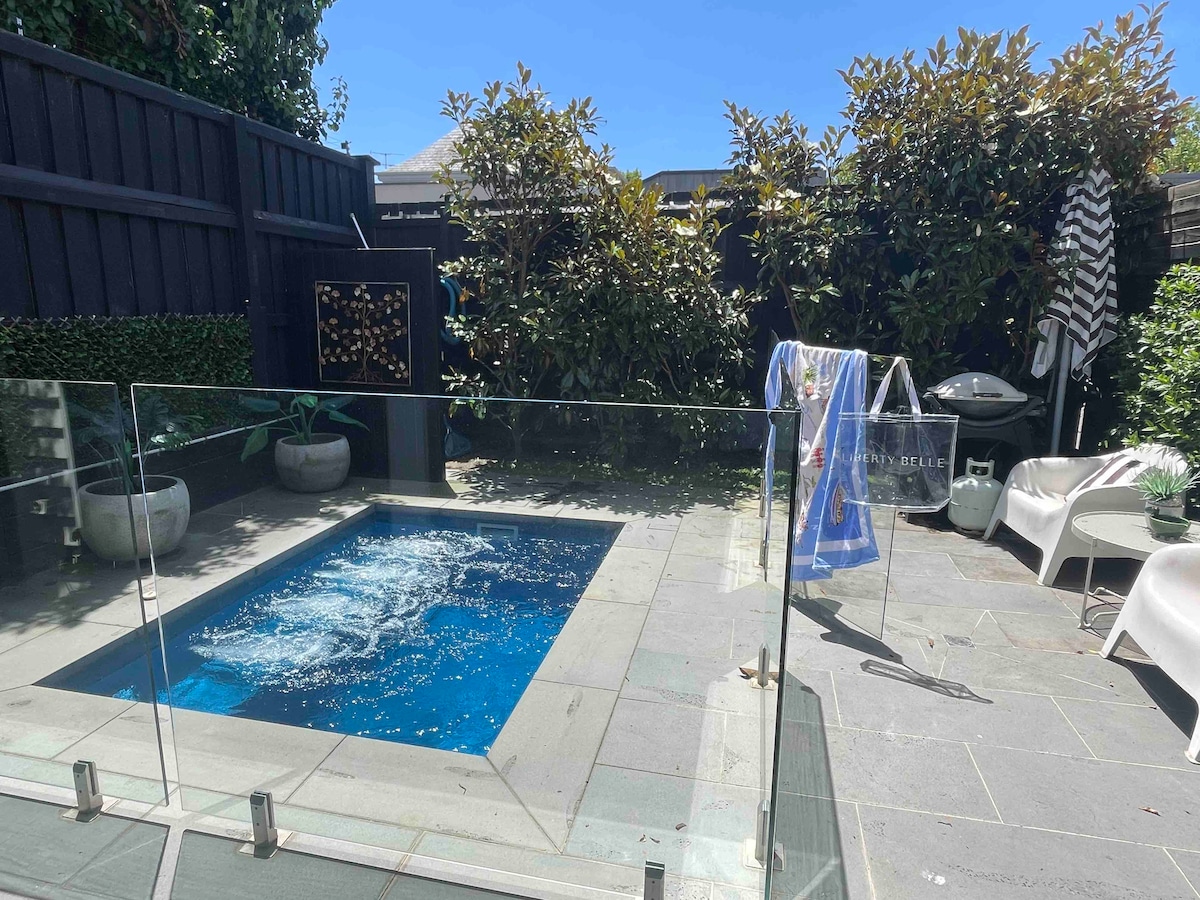 A small outdoor area features a crystalline blue plunge pool surrounded by a stone patio. Lush greenery is visible against the dark wooden fence. There are two lounge chairs positioned nearby, and a clothing rack holds wet towels and swimsuits, adding a casual touch to the space.