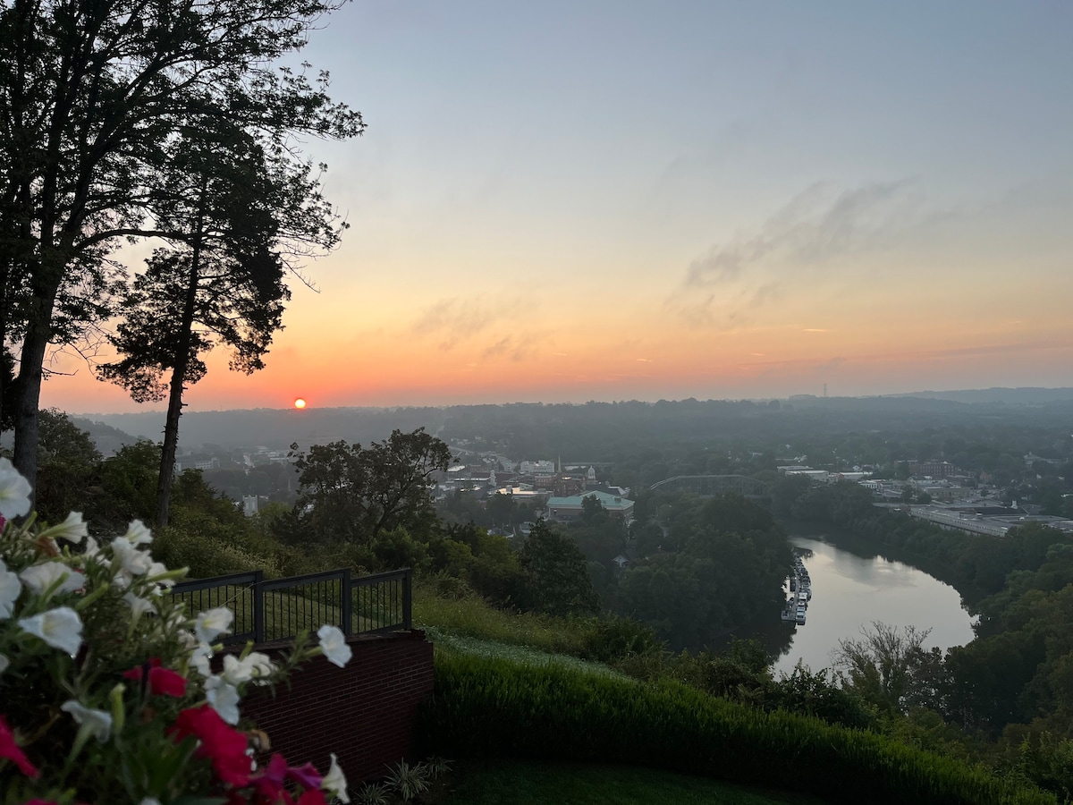 A scenic view captures the sunrise over Frankfort, featuring the Kentucky River winding through the landscape. The horizon is painted with soft hues of orange and pink, while clusters of flowers in the foreground add a touch of color to the serene setting.