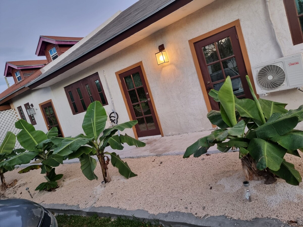 The exterior of the building is characterized by a textured white facade and a gently sloped roof. Lush banana plants lined with decorative stones create a welcoming entrance, while large windows and a warm light fixture enhance the inviting atmosphere.