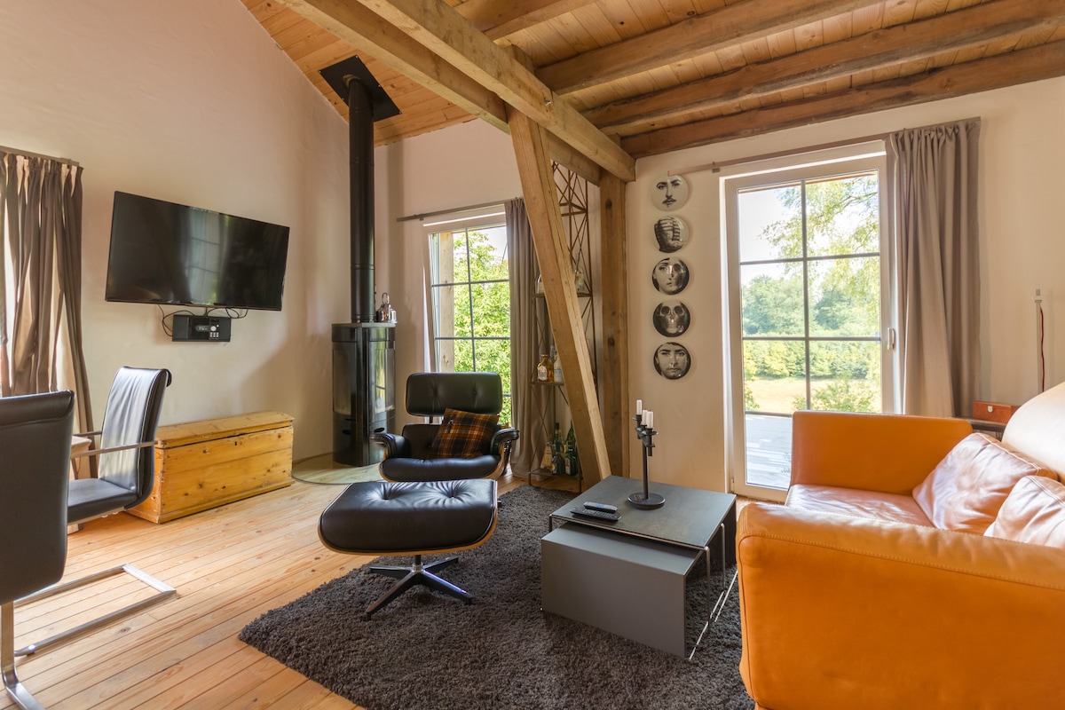 A living area features wooden beams and a cozy atmosphere. A stylish orange sofa is paired with a black lounge chair, positioned near a sleek coffee table. Natural light fills the space through large windows, revealing views of the surrounding greenery. A modern television is mounted on the wall.