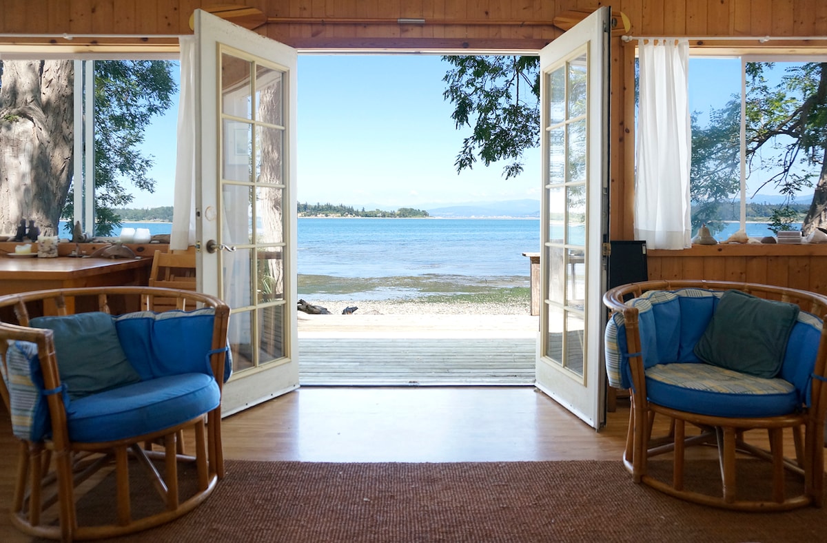French doors frame a scenic view of the shoreline and water. Two wicker chairs with blue cushions are positioned inside, enhancing the openness of the space. Natural light filters through the doors, illuminating the wooden interior and creating a serene atmosphere.
