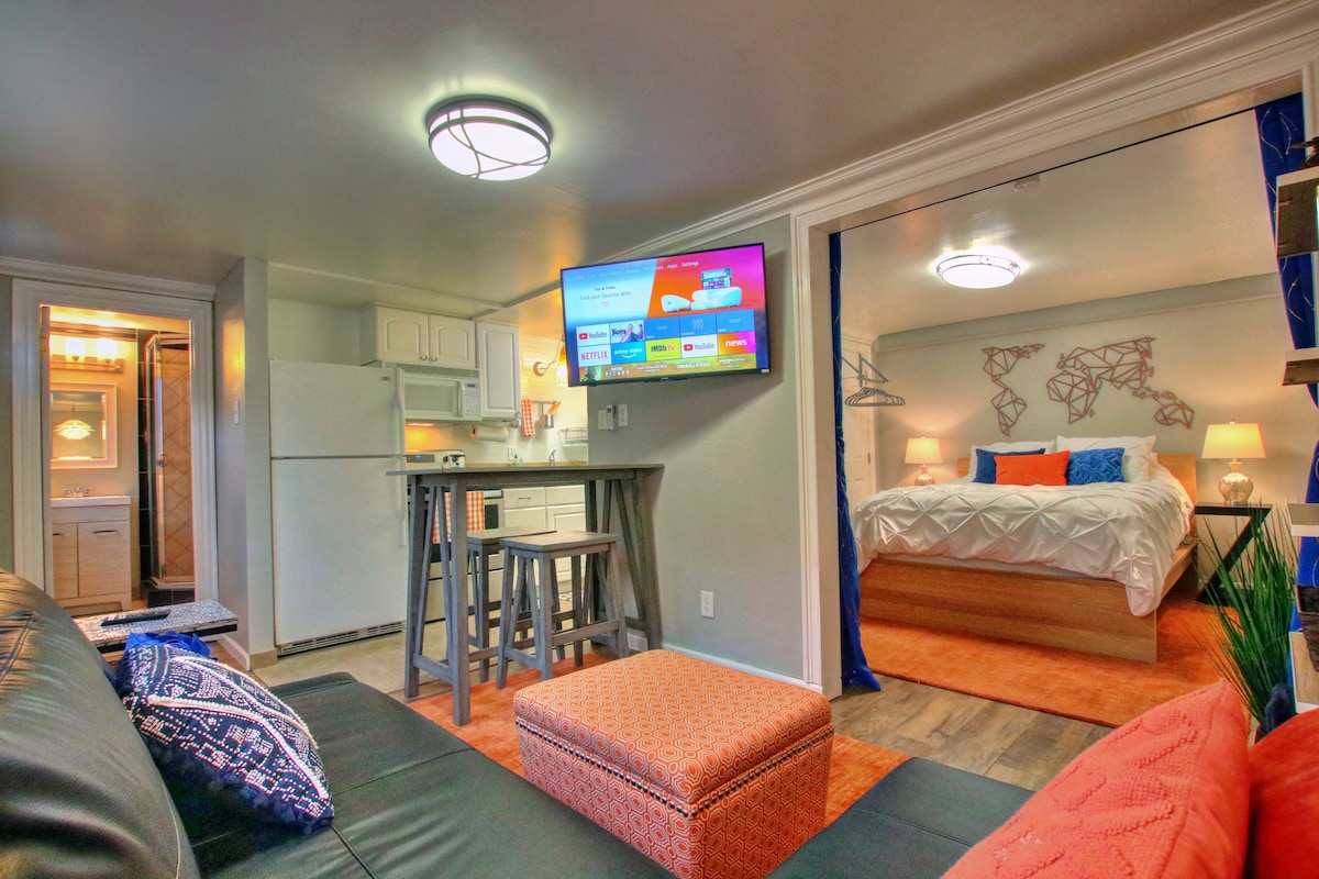 The suite features a comfortable living area with a black sofa, an orange pouf, and a wall-mounted TV. A small dining table and kitchenette are seen in the background, while a bedroom with a king-sized bed is visible through an archway. The bathroom door is also present, highlighting the suite's layout.