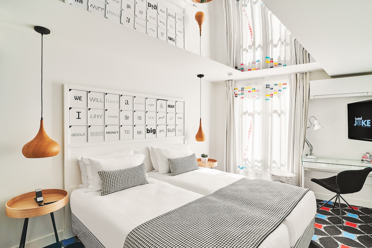 A twin room features two neatly arranged beds with checkered bed linens. A modern art piece hangs above the beds, while large windows allow natural light to fill the space, accented by colorful decorative elements. A desk is positioned beside the beds, with stylish lighting overhead.