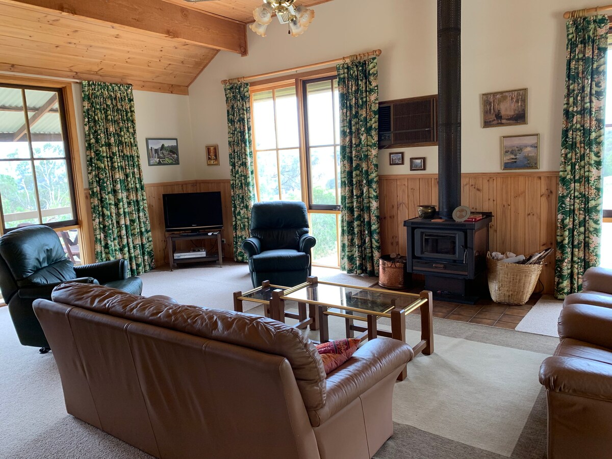 A spacious living area features large windows allowing natural light to fill the room. Comfortable leather sofas are arranged around a coffee table, while a wood stove provides warmth. A television and decorative elements contribute to a relaxed ambiance, framed by nature views.