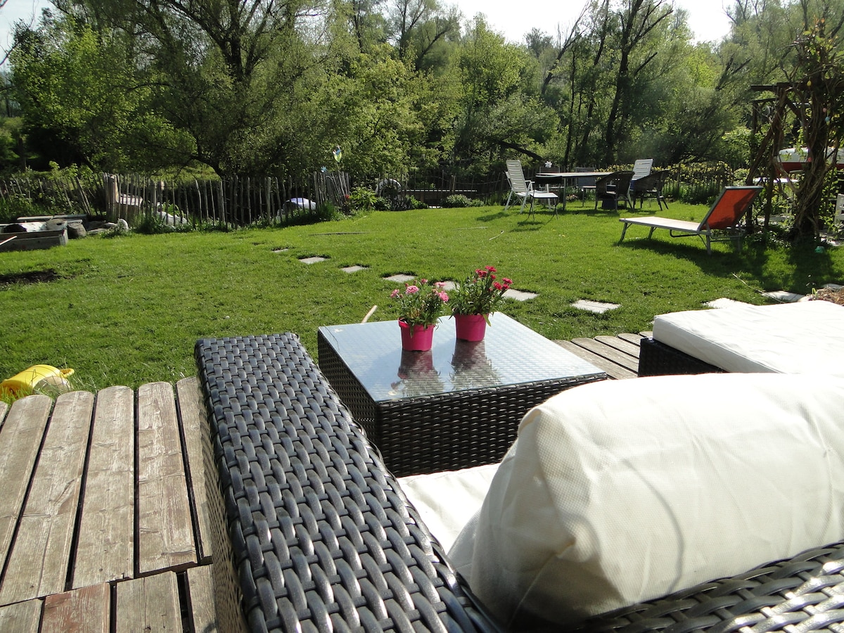 The outdoor seating area features a wicker lounge set topped with light cushions, situated on a wooden deck. A glass table is placed centrally, surrounded by lush green grass and vibrant potted flowers. In the background, various seating options are visible among trees.